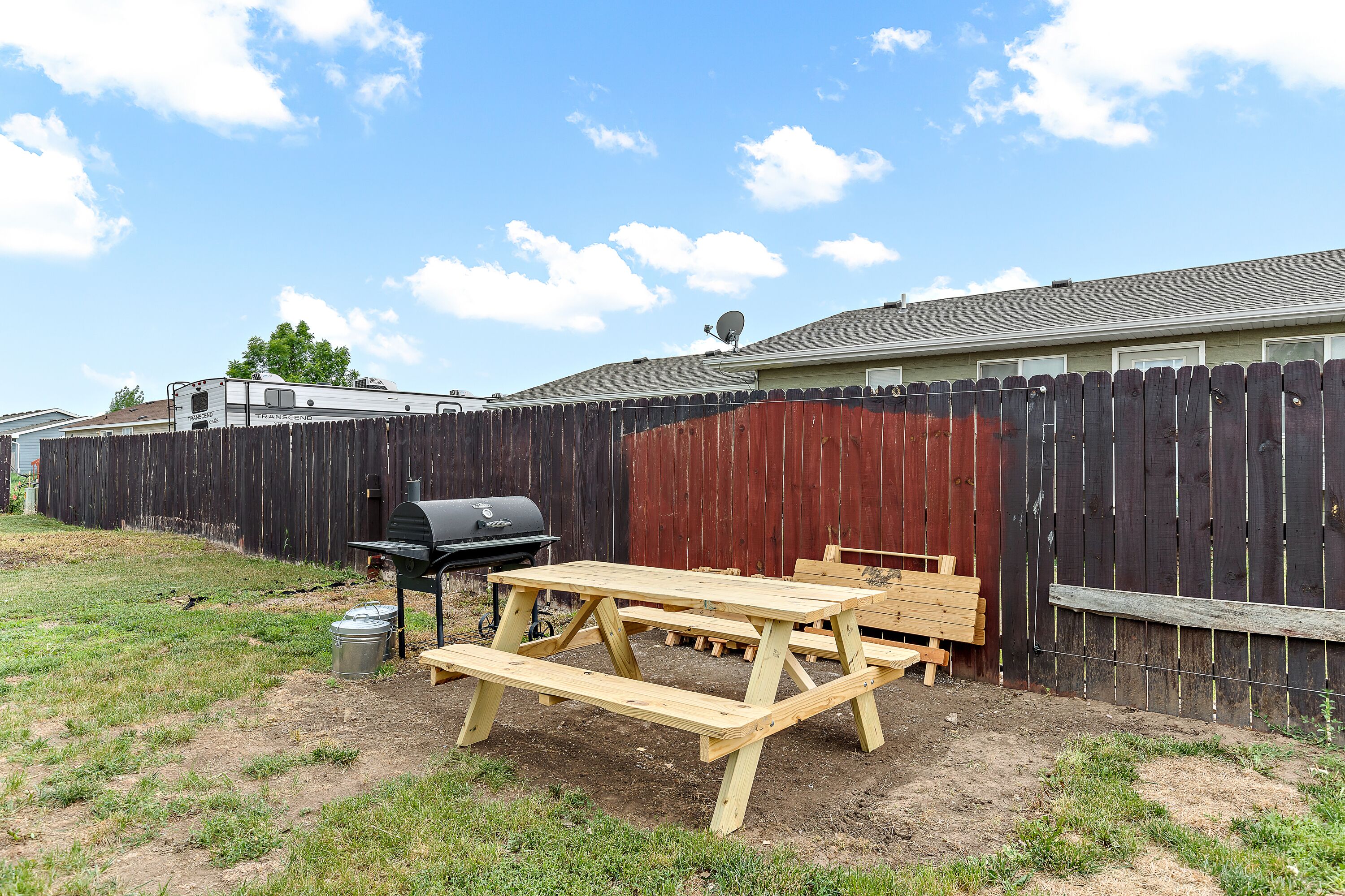 A backyard with a wooden picnic table, a grill, and a fenced area.