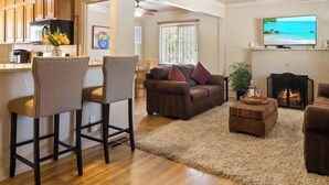 A living room with a sofa, two bar stools, a fireplace, a television, and a kitchen area with wooden cabinets and a dining table. - The Dartmouth House S (Albuquerque)