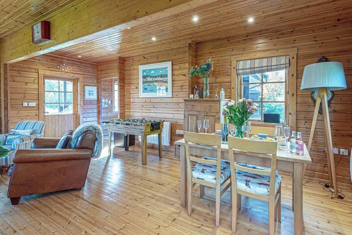 A cozy cabin interior with wooden walls, a foosball table, a dining area with chairs and a table set for two, and a sofa in the living area.