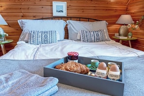 A cozy bedroom with a wooden wall, a bed with patterned pillows, a bedside table with a lamp, and a tray with food and drinks. - Scandi Cabin in Heart of Anglesey with Parking (Anglesey)