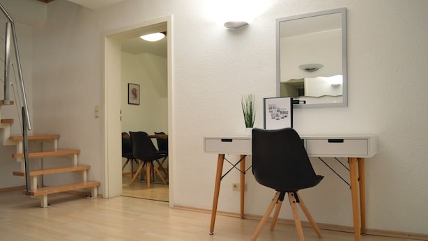 A modern room with a desk, chair, and a mirror. There is a staircase on the left and a door leading to another room.