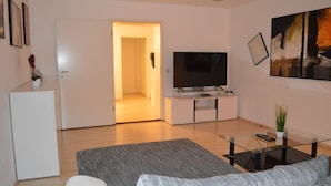 A modern living room with a flat-screen TV, a glass coffee table, and a grey sofa.