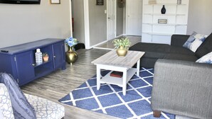 Een woonkamer met een blauw-wit gestreept tapijt, een donkerblauwe kast, een witte salontafel met een gouden vaas, een grijze bank en een grijze fauteuil.