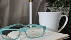 Een bril op een open boek, een witte mok en een pen op een oppervlak.