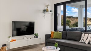 Un soggiorno moderno con una grande televisione a schermo piatto, un divano grigio con cuscini, un tavolino rotondo e una finestra che si affaccia su un paesaggio collinare.
