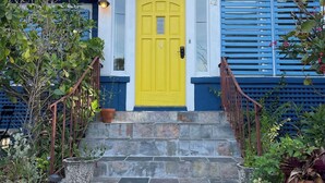 Uma casa com uma porta amarela vibrante, janelas com persianas azuis e uma escadaria de pedra que leva à entrada.