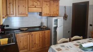 A kitchen with wooden cabinets, a stainless steel sink, a white refrigerator, and a dining table with a patterned tablecloth. - La Via Antiga - Apartment # 1 (Torre di Mosto)