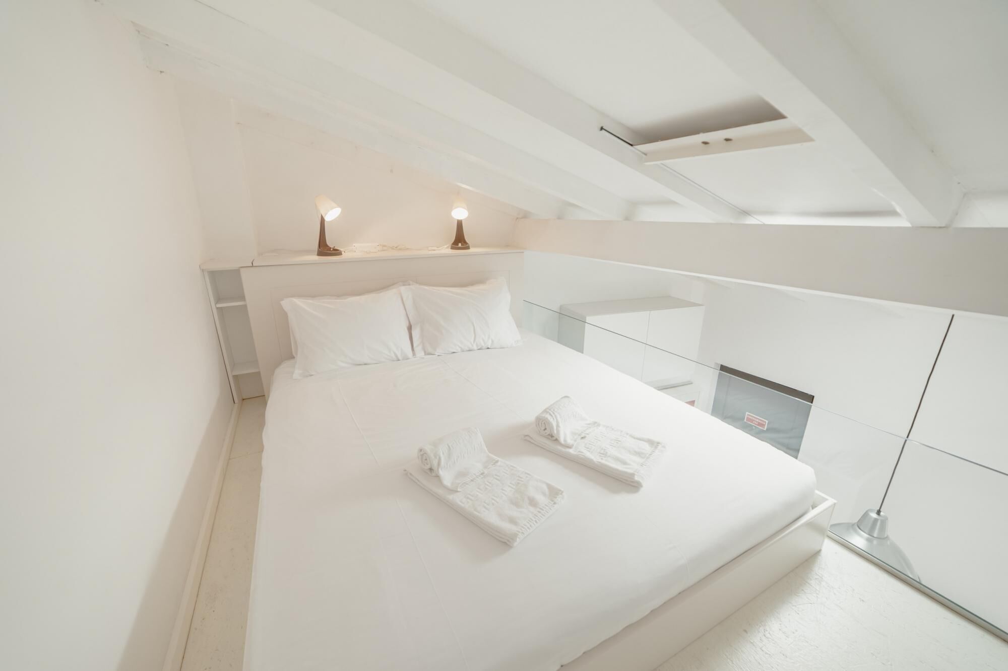 Ein modernes Schlafzimmer in Weiß mit einem großen Bett, zwei Nachttischlampen und einem Glasgeländer mit Blick auf einen Balkon.