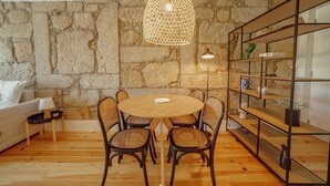 Ein gemütlicher Essbereich mit einem runden Holztisch, Korbstühlen, einer Steinwand und einem Metallregal mit diversen Gegenständen.