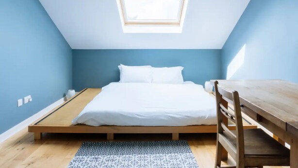 A room with a sloped ceiling, a skylight, a bed with white bedding, a wooden table, and a patterned rug. - Stylish 3 Bedroom Apartment Near Hollyrood Park (Edinburgh)