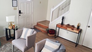 Un salon comprenant deux fauteuils, une petite table avec une lampe, un escalier et une console ornée d’objets décoratifs.
