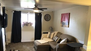 Un salon avec un canapé, un chien allongé sur le sol, un ventilateur de plafond et une fenêtre avec des rideaux.
