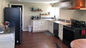 Une cuisine moderne avec des armoires blanches, un réfrigérateur noir, un four à micro-ondes, une cuisinière et un évier. On trouve des étagères en bois au-dessus du plan de travail et une petite poubelle à gauche.