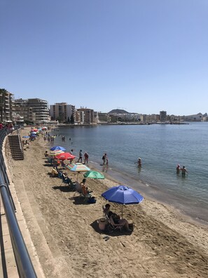 A beach with rows of umbrellas and chairs, a clear sky, and buildings in the background.