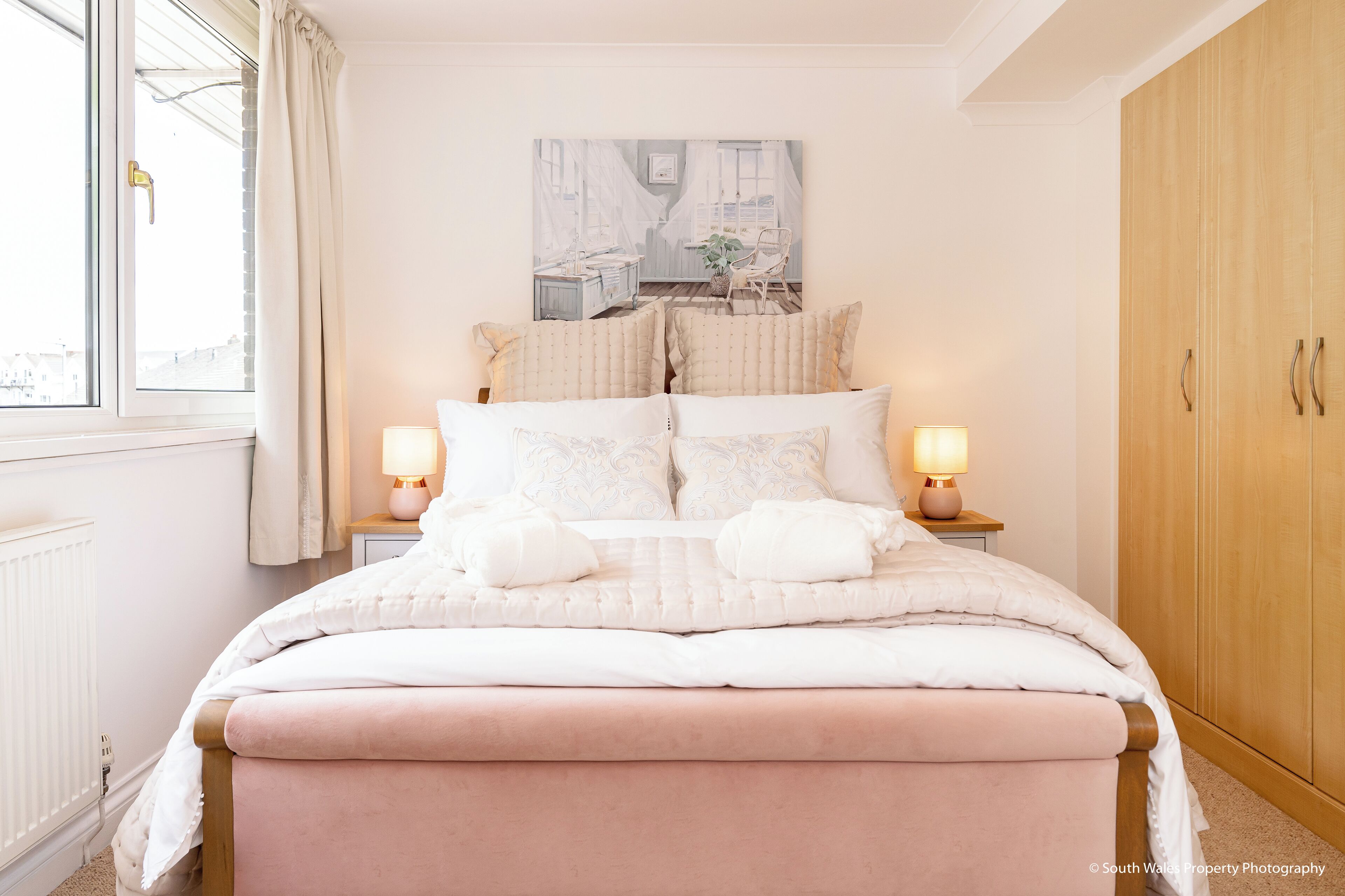 Un dormitorio con cama, lámparas de noche, una ventana con cortinas, un armario de madera y un cuadro en la pared.