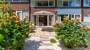 Una casa de dos pisos con un patio que incluye una mesa y sillas, rodeado de plantas en flor.