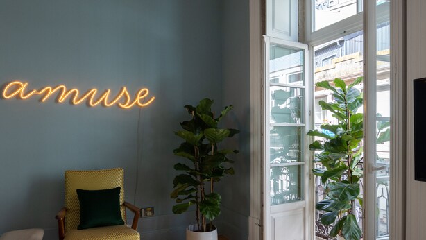 A room with a blue wall, a neon sign reading "amuse", a yellow armchair, a large potted plant, and a view through a glass door. - Almadina Smart Luxury (Porto)