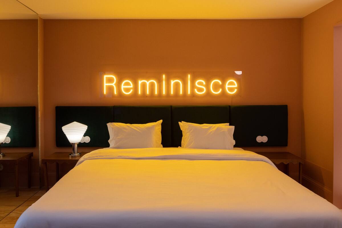 Hotellihuone, jossa on sänky, kaksi yöpöytää, yölamppu ja seinällä oleva neonvalomainos.