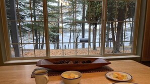 A breakfast table with plates of scrambled eggs, toast, and fruit, set against a window with a view of a forest and a body of water. - Beautiful Lake View - Townhome (Saint Germain)
