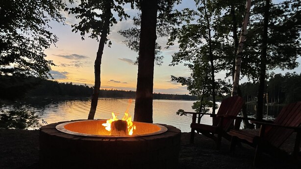 A campfire in a circular fire pit with Adirondack chairs nearby, overlooking a lake at sunset. - Beautiful Lake View - Townhome (Saint Germain)