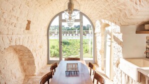 Interior rústico em pedra, com mesa e cadeiras de madeira, uma grande janela em arco e vista para um jardim.