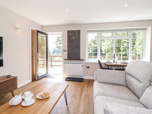 A modern living room with a sofa, a wooden coffee table with teapots and cookies, a dining area with a table and chairs, and a large window with a view of greenery. - Ginger &amp; Pickles (Windermere)