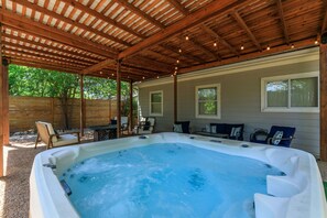 A hot tub under a wooden pergola with seating and a view of a house. - The Emerald Door | Hot Tub | Fire Pit (Fredericksburg)