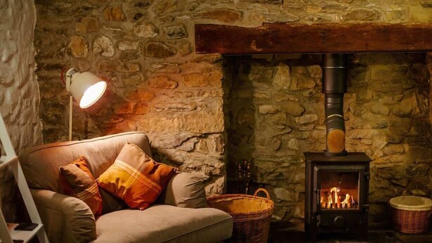 A cozy room with a stone wall, a sofa with cushions, a standing lamp, a fireplace with a wood-burning stove, and a wicker basket. - St Davids Hideaway with Roll-top Bath & Log Burner (St Davids)