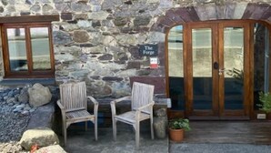 A stone building with two wooden-framed windows and a glass door. There are two wooden chairs and a small table in front of the building. - St Davids Hideaway with Roll-top Bath & Log Burner (St Davids)