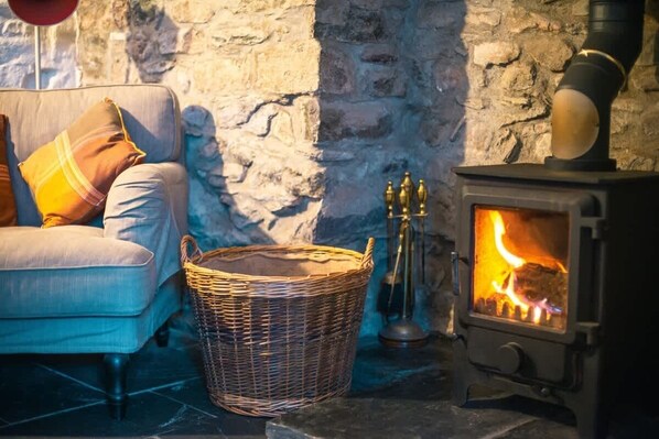 Une pièce chaleureuse avec un mur en pierre, un canapé avec des coussins, un panier en osier et un poêle à bois.