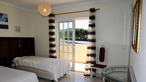 Habitación de hotel con dos camas, una mesa de vidrio, una silla, un sillón de madera, una lámpara colgante y vistas a través de la puerta.