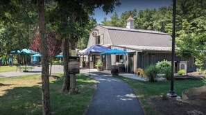 A building with a covered outdoor seating area, a pathway leading to the entrance, and a garden with trees and shrubs.