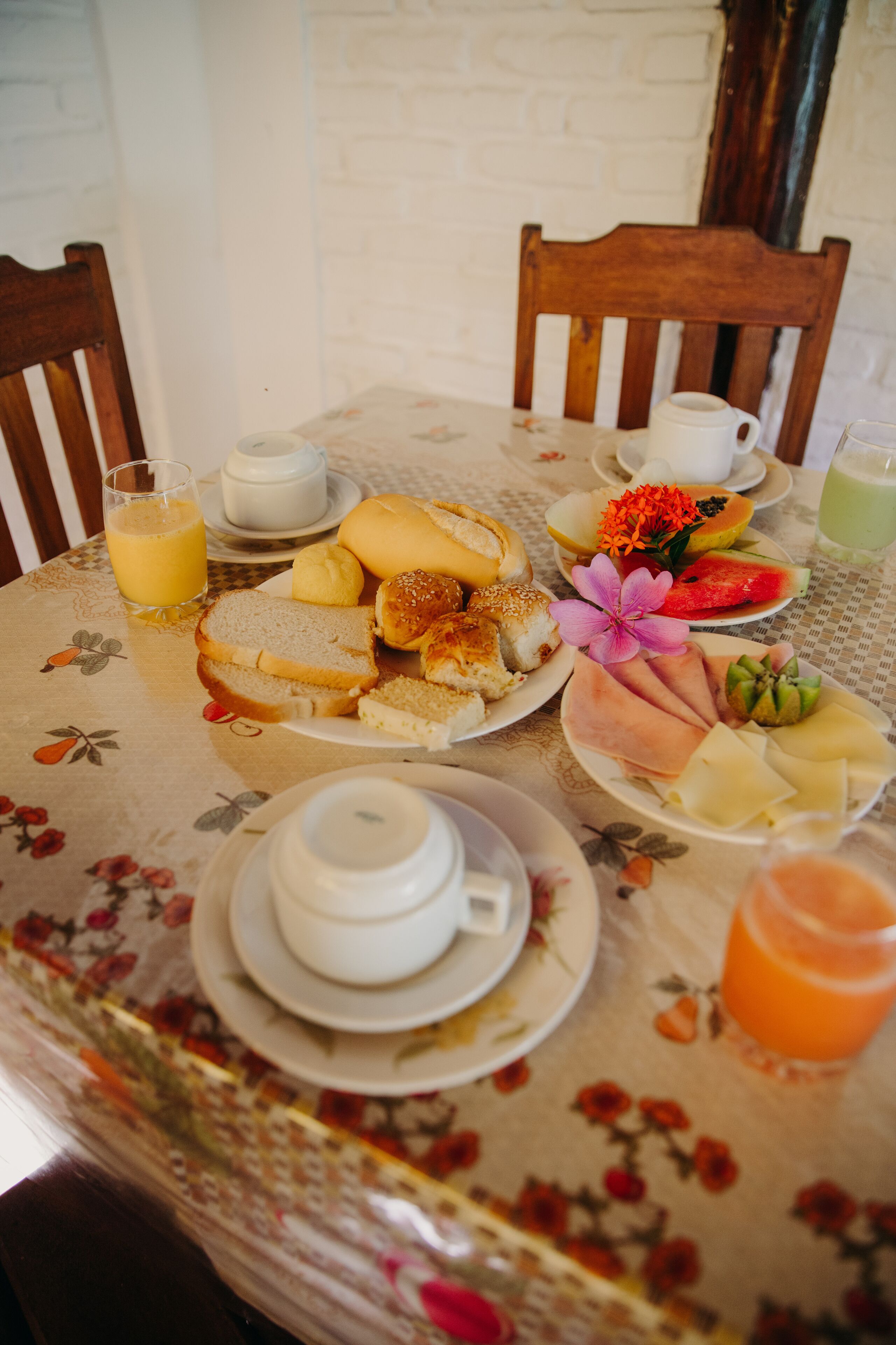 Mesa posta para o pequeno-almoço com pão, ovos e fruta, com toalha de mesa florida.