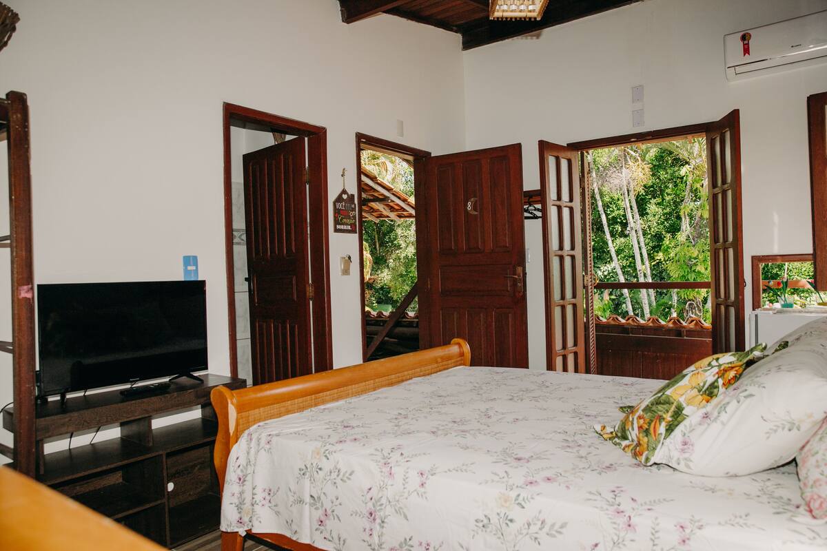 Un dormitorio con cama, televisor sobre un mueble de madera y puerta que da a un balcón con vistas a la vegetación.