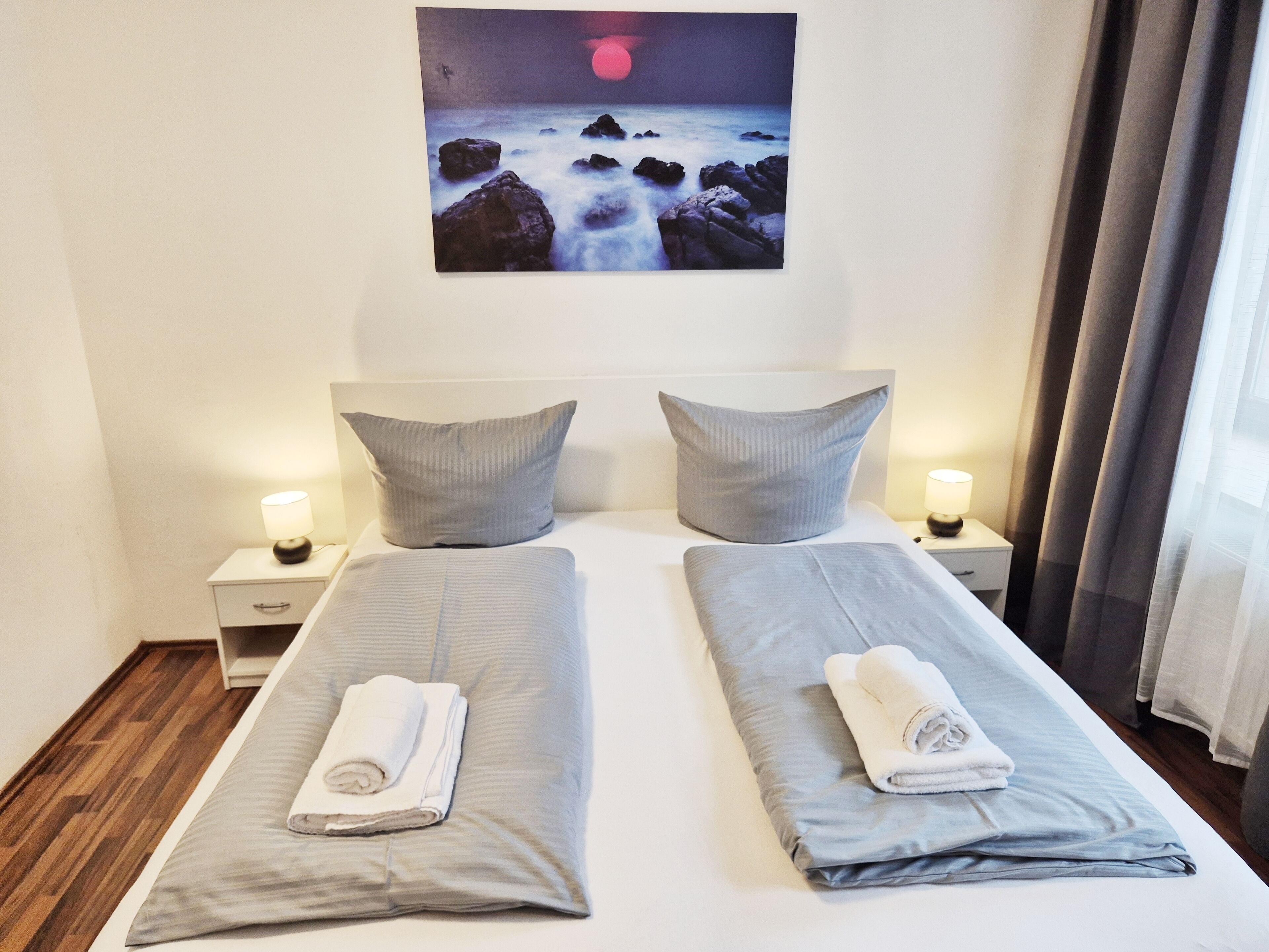 A double bed with grey linens and two pillows, a nightstand with a lamp, and a painting of a rocky coastline above the bed.