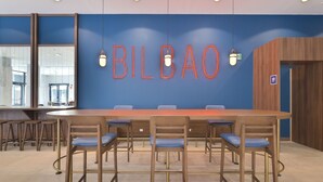 Une salle de conférence avec une longue table et six chaises, un mur bleu sur lequel le nom « BILBAO » est écrit en lettres néon, et un panneau en bois sur le côté droit.