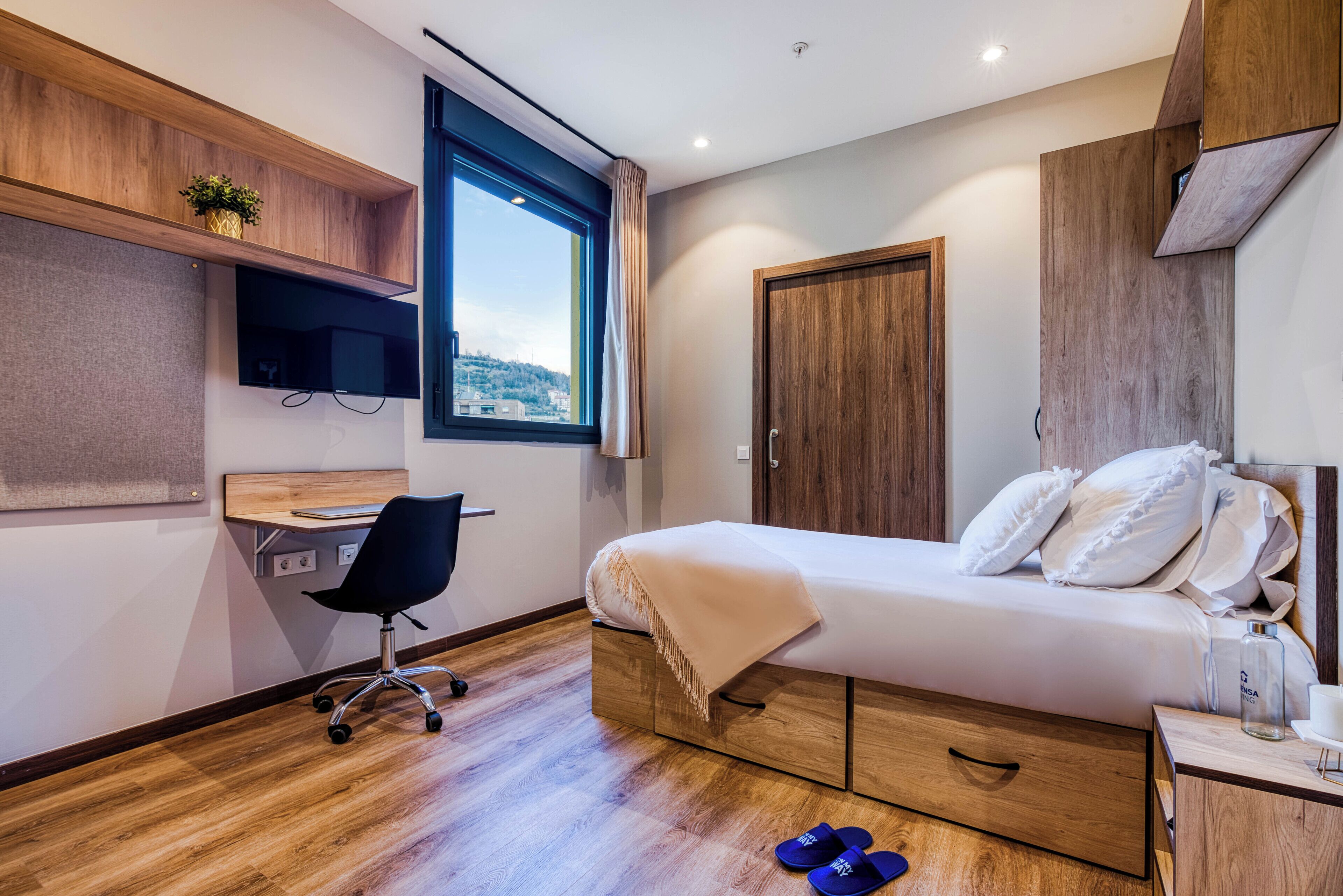 A modern bedroom with a bed, desk, chair, and a window.