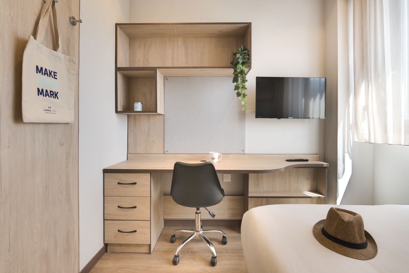 A modern hotel room with a wooden desk, a comfortable chair, a bed with a hat, a TV, and a plant.