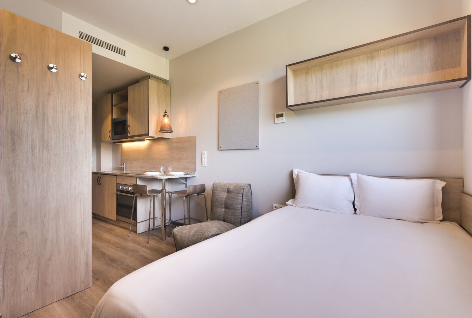 Une chambre d’hôtel moderne dotée d’un grand lit, d’un bureau avec deux chaises, d’une kitchenette et d’une armoire en bois.