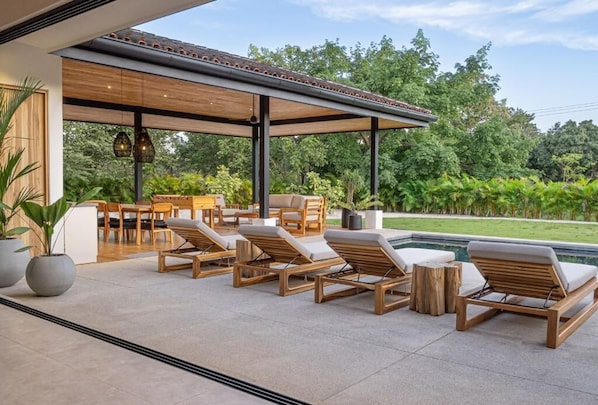 A covered outdoor seating area with lounge chairs and a dining table, overlooking a swimming pool and lush greenery.