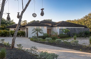 A modern house with a stone facade, a tiled roof, and a landscaped garden with various plants and a palm tree.
