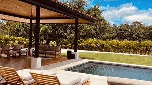 A poolside area with wooden loungers, a covered seating area, and a view of a lush garden and pool. - Casa Zuri Luxury Villa + Guest House & Beach Club (Tamarindo)