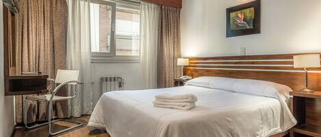 A hotel room with a large bed, a desk with a chair, a bedside table with a lamp, and a window with curtains.