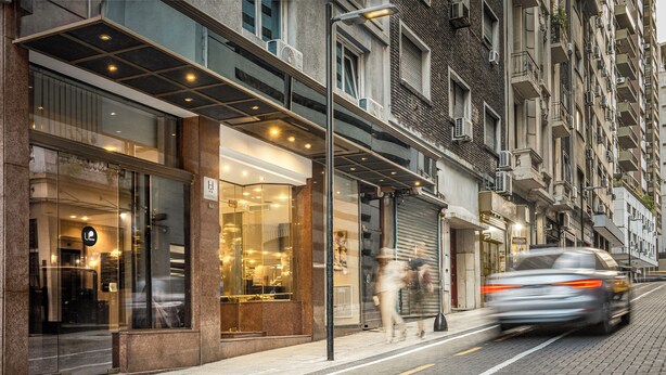 En moderne hotelfacade med store glasvinduer, en overdækket indgang og udsigt til gaden med en forbipasserende bil.