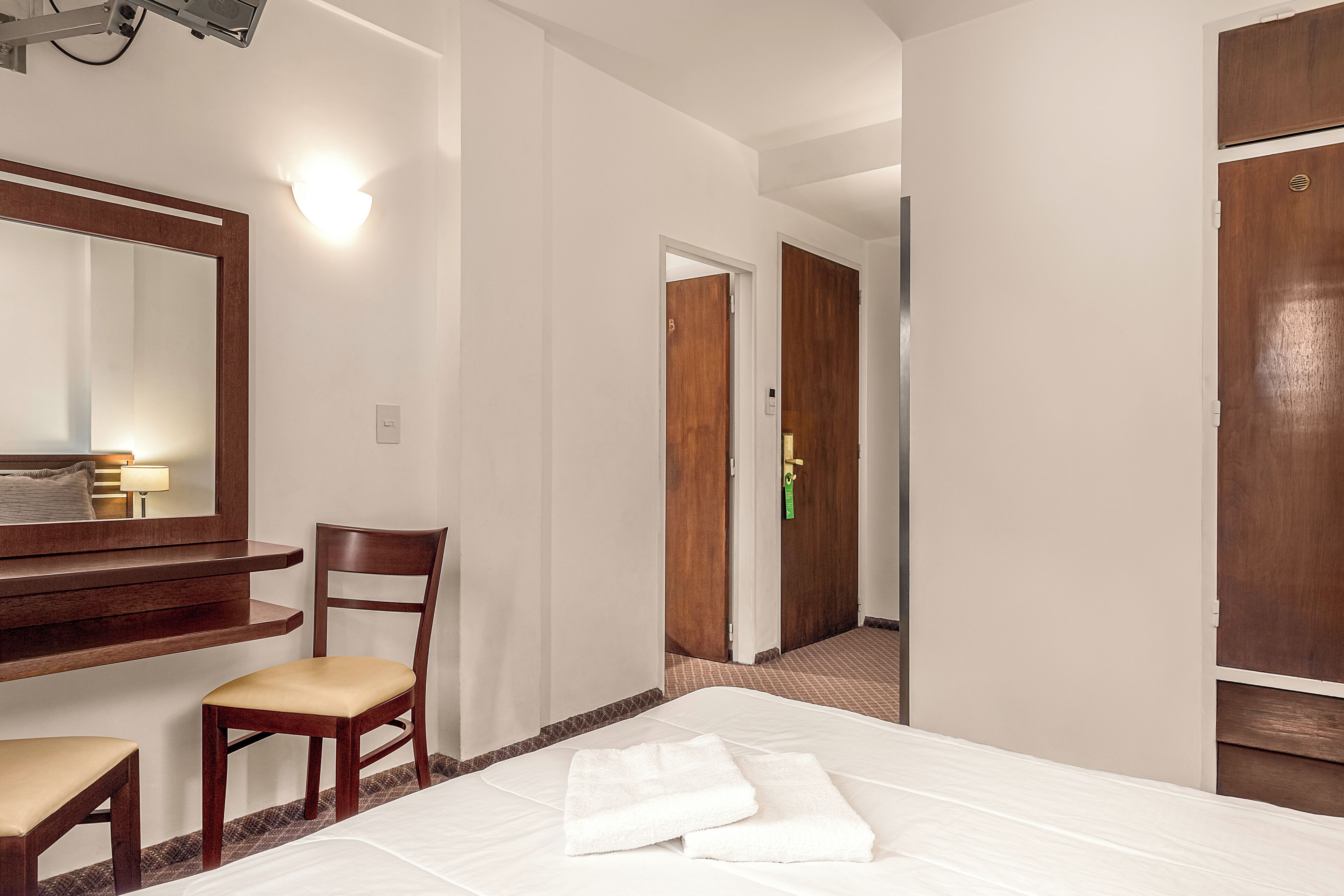 A hotel room with a bed, a chair, a desk, a mirror, and a door.