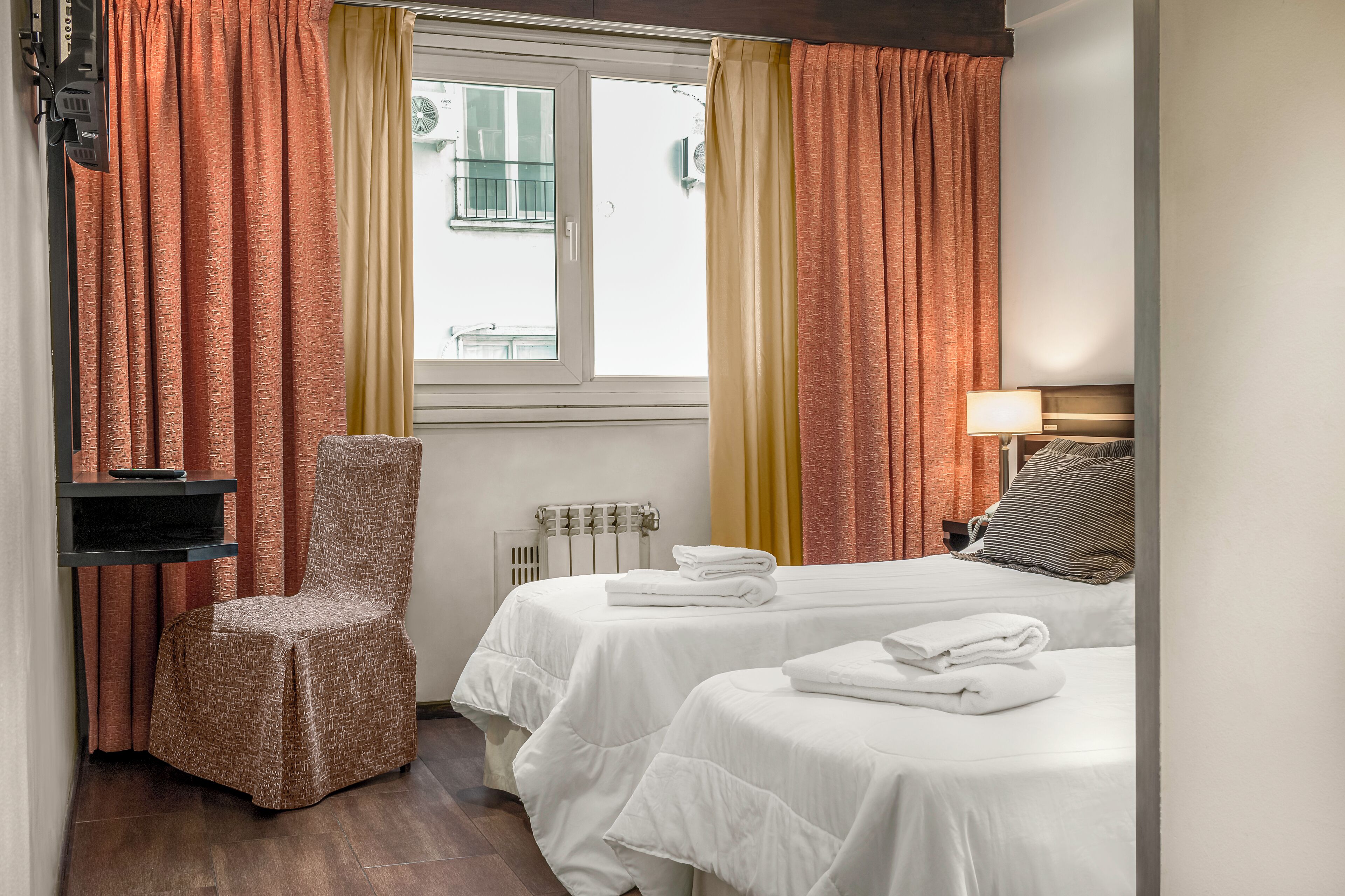 A hotel room with two beds, a chair, a radiator, and a window with curtains.