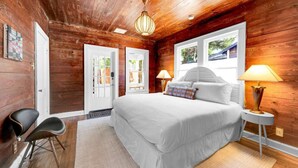 A bedroom with a large bed, two bedside lamps, a wooden ceiling, and a view through a window.