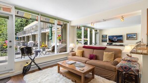 Un salón con sofá, mesa de centro y televisor. Amplias ventanas ofrecen vista a un edificio y follaje exterior.