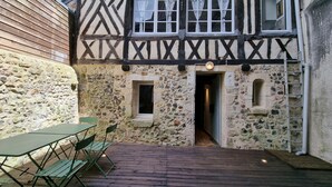 Un bâtiment traditionnel à colombages, doté d’une façade en pierre, d’une terrasse en bois avec une table et des chaises, et d’une petite porte donnant accès à un espace intérieur.