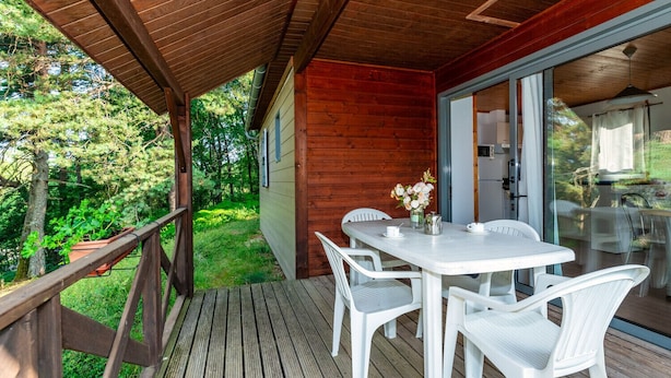 Una terraza de madera con una mesa y sillas, un ventanal de vidrio que da a una cocina, y vista a un área boscosa.